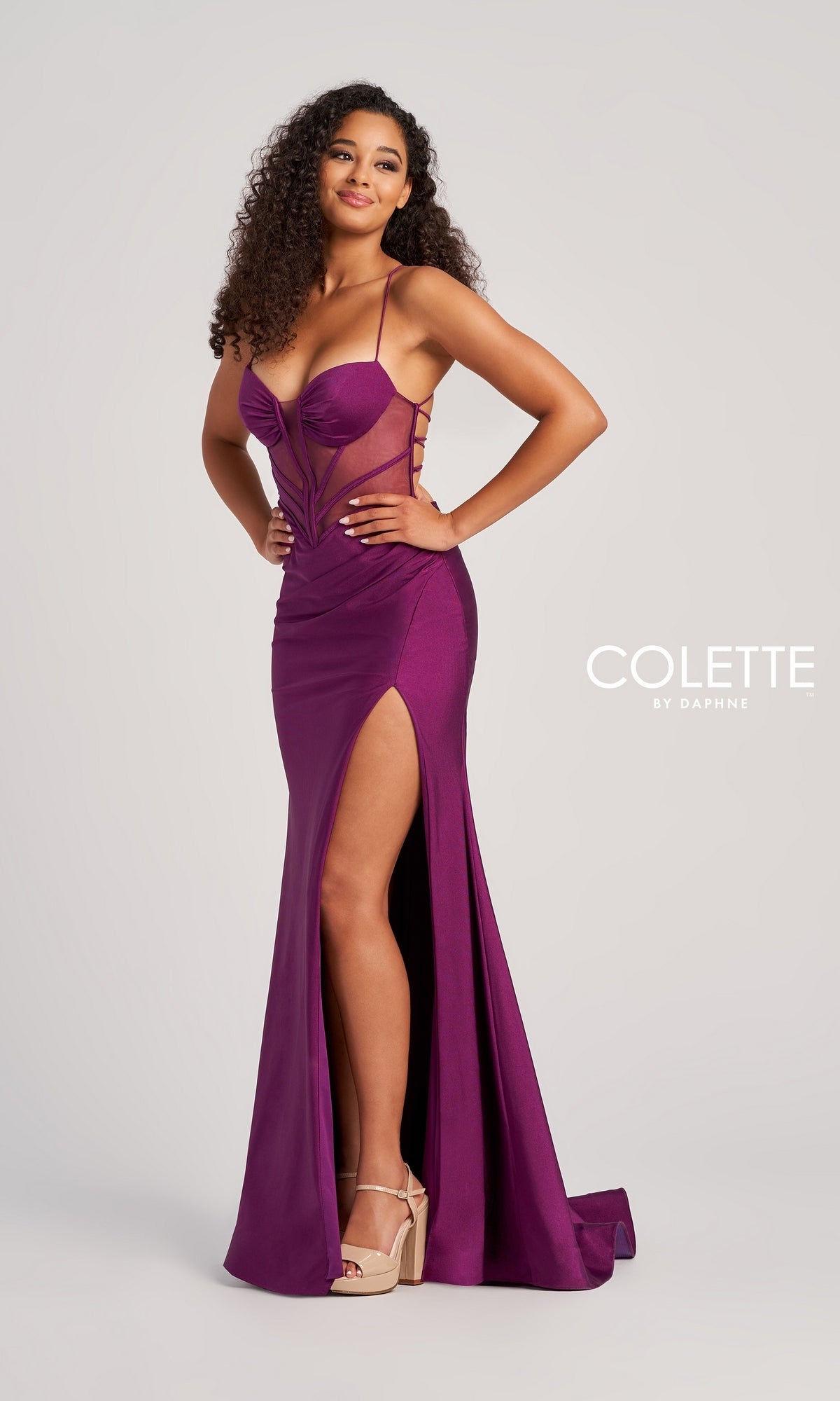 A woman poses against a plain background in the Backless Colette Long Designer Prom Dress CL5140, featuring a purple hue, floor-length cut with high slit, sheer paneling, spaghetti straps, and platform heels.